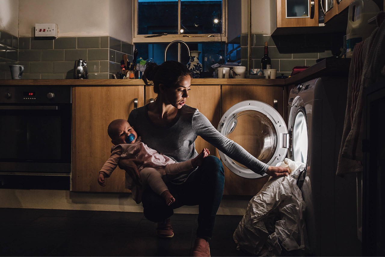 Frau trägt schreiendes Baby auf dem Arm und räumt die Waschmaschine aus