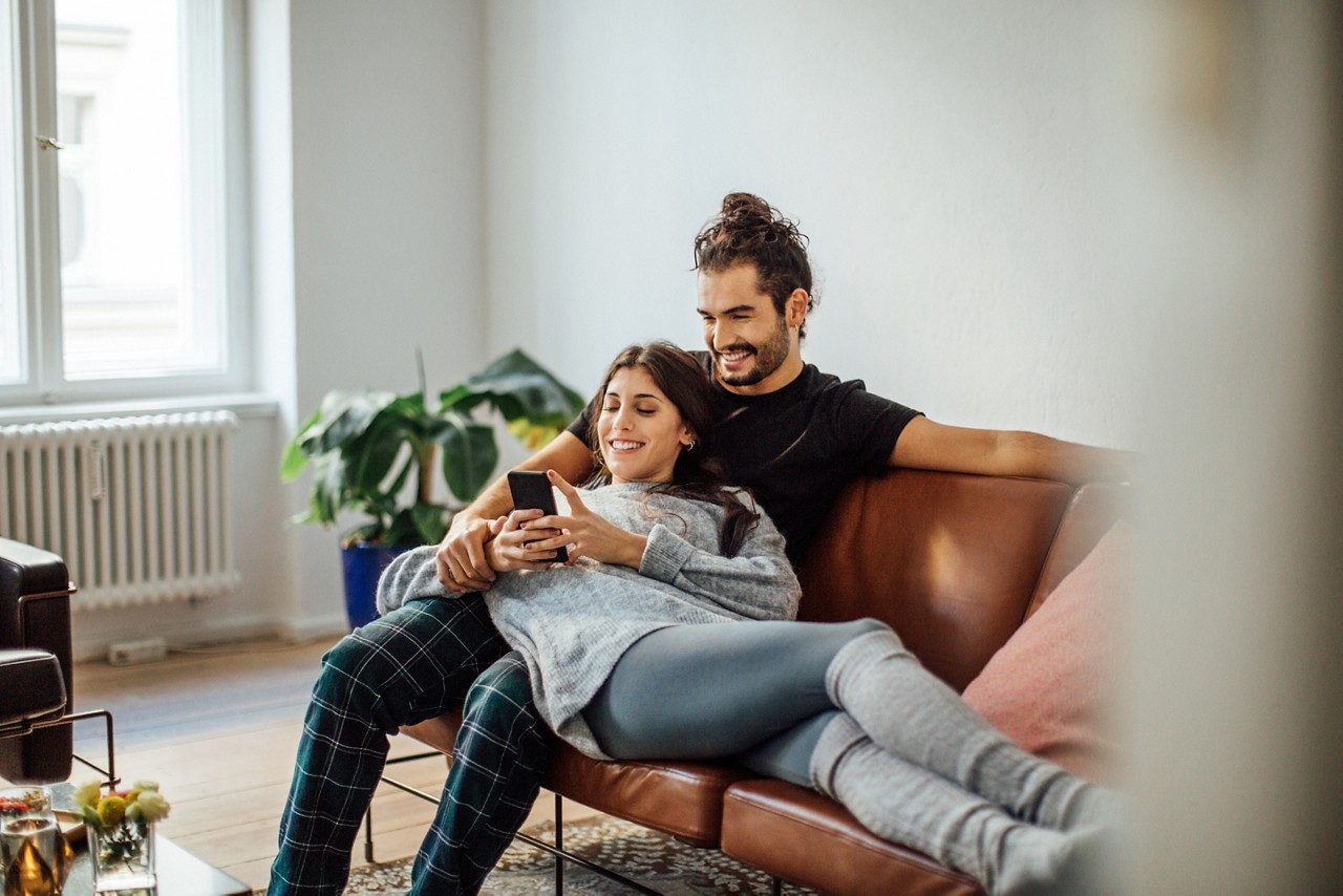 Smiling young woman using mobile phone while reclining on boyfriend sitting on sofa at home