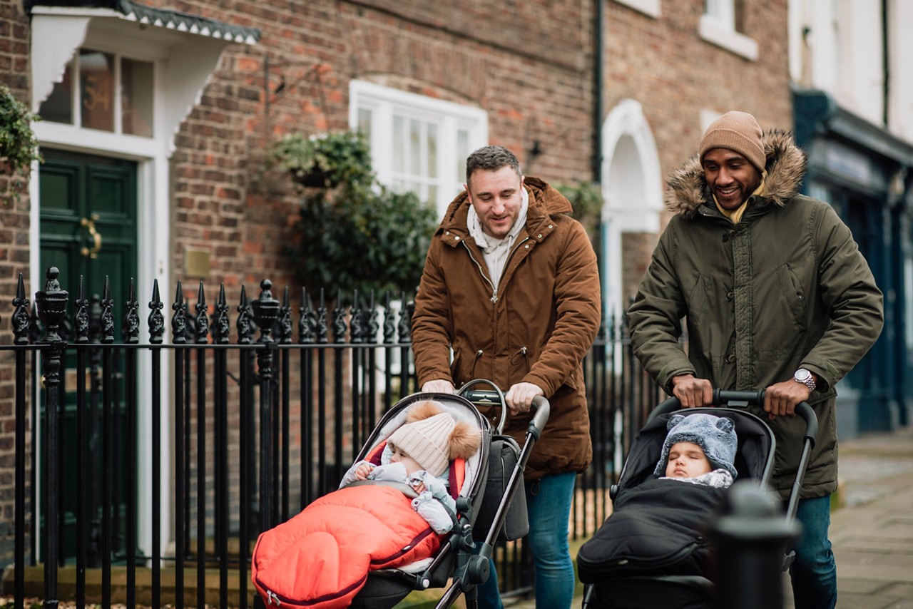 Two male friends are out in Tynemouth, North East UK. They are walking on a sidewalk and pushing their baby sons in strollers. They are wearing warm clothing.