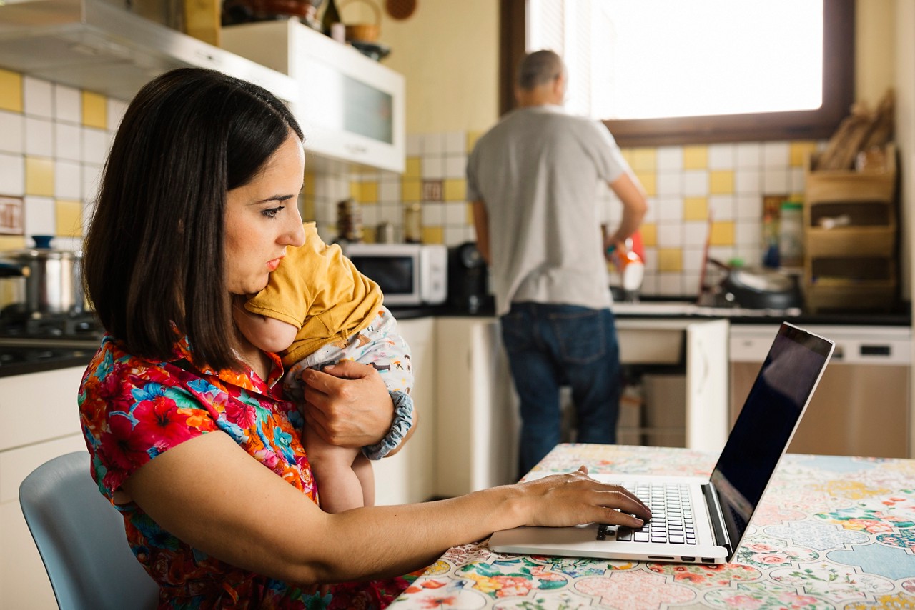 Young woman in a kitchen working on her laptop while holding her baby on her arms