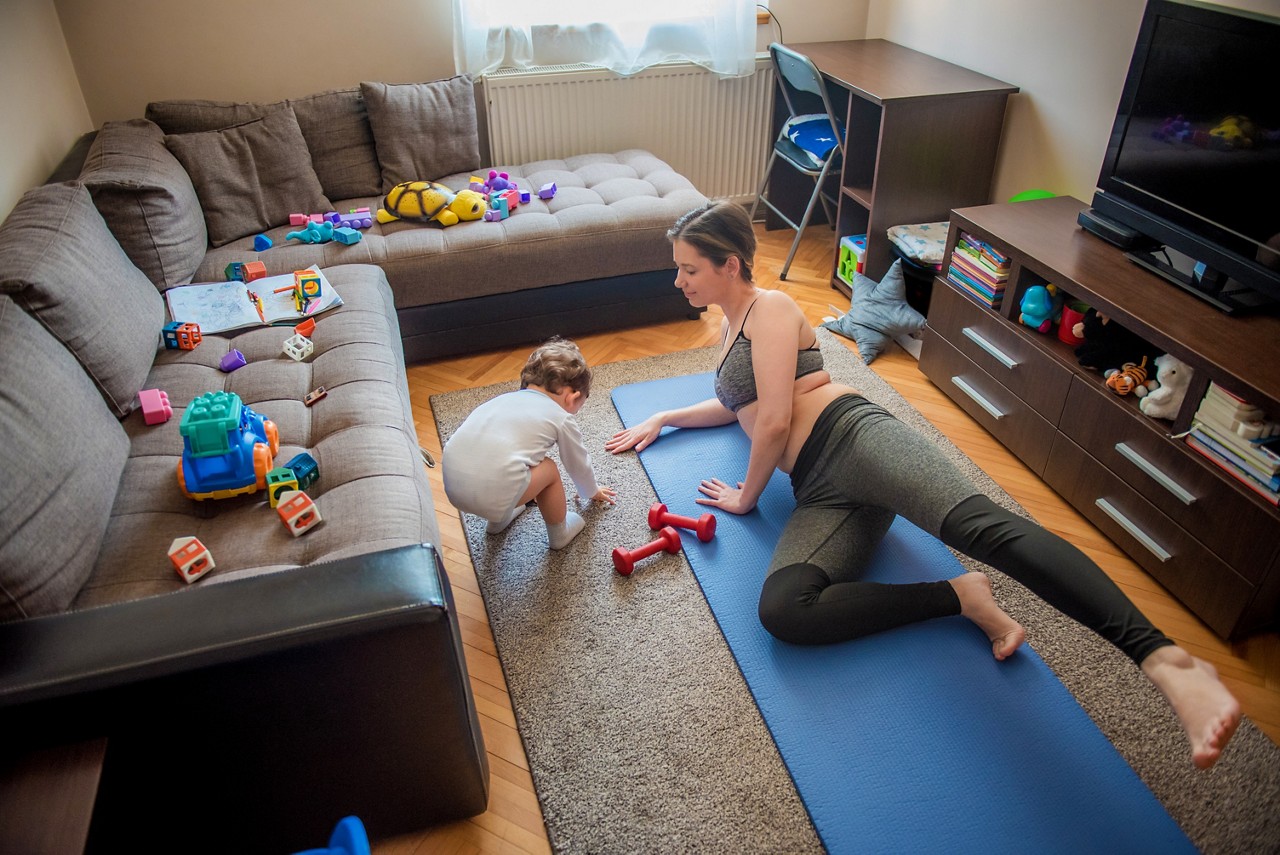 Young mother, exercising at home while her toddler son happily playing around her with toys. Woman staying in shape after pregnancy, during coronavirus, covid 19 quarantine. Young family spending time together living healthy.