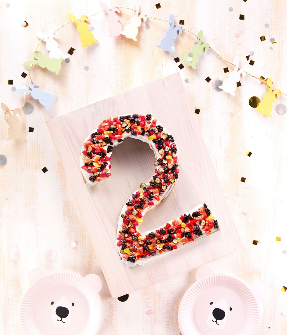 Un g�teau festif en forme de chiffre 2, decore de fruits et de bonbons colores. Le decor comprend des guirlandes de lapins pastel et des confettis sur une surface en bois clair. Deux assiettes sur le theme des ours ajoutent une touche fantaisiste a la fete.