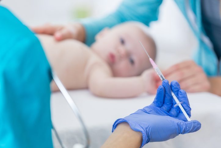 A female healthcare professional holds a syringe as she prepares to give a baby girl a vaccine during the child's well check appointment. The healthcare professional is wearing a protective glove. Focus is on the woman's hand holding the needle. The baby is blurred in the background.