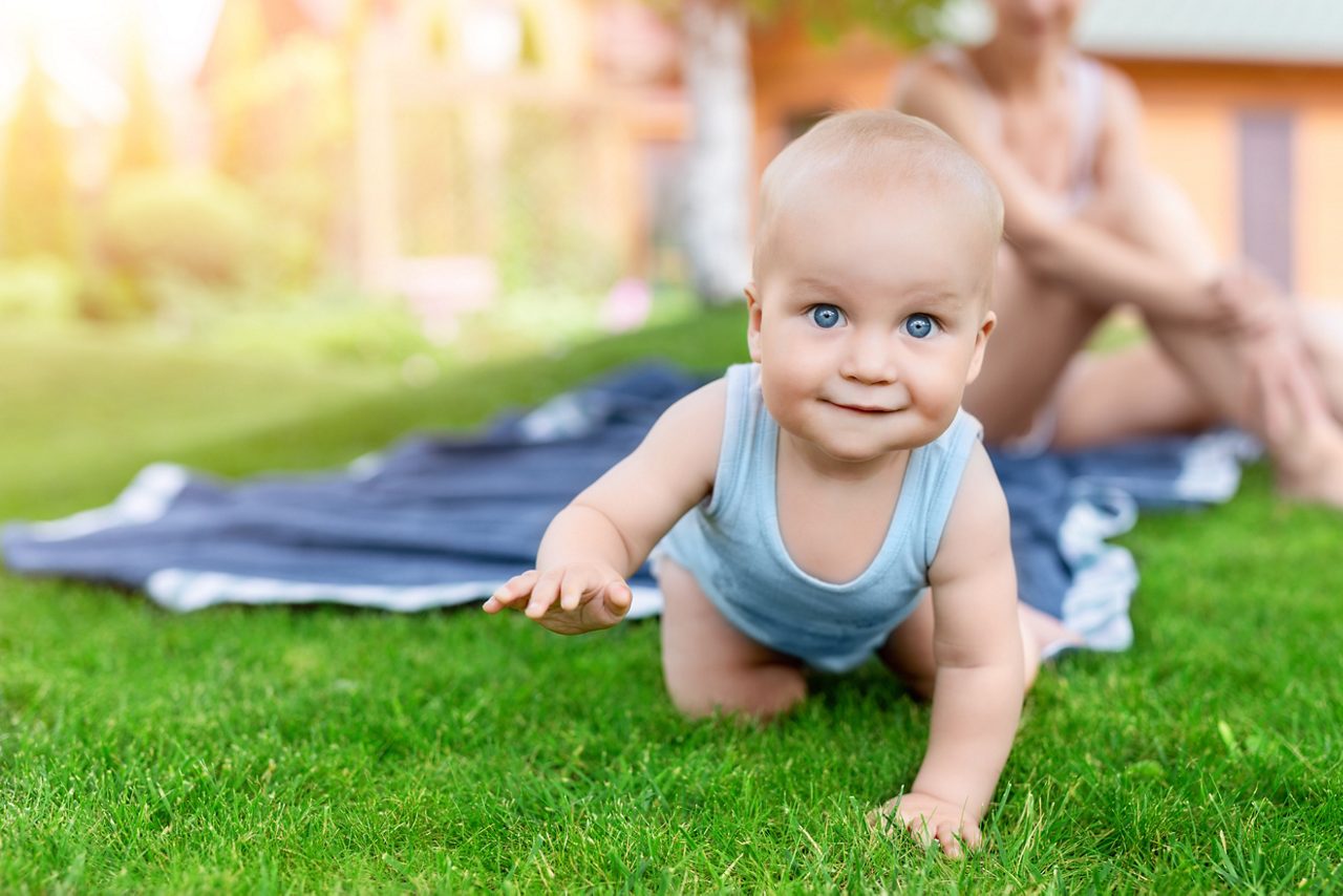 Portrait of cute little caucasian boy having fun in garden with mother. Child crawling on green grass lawn during walk with mom in yard. Happy childhood and baby healthcare.