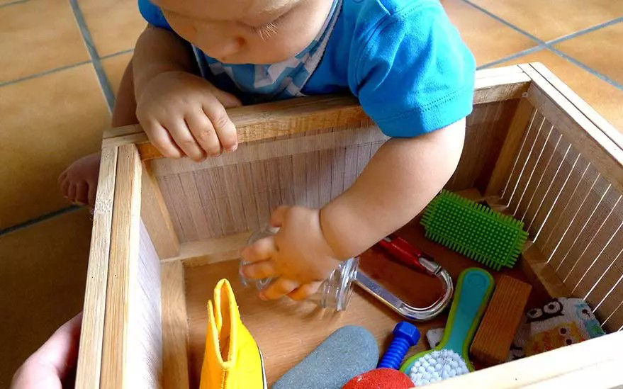 On voit un jeune bebe vetu d'une chemise bleue plonger la main dans une bo�te en bois remplie d'objets colores. Le decor semble etre a l'interieur, sur un sol carrele. La bo�te contient des articles tels qu'une chaussure jaune, une bouteille en verre, des brosses et d'autres petits objets. Le bebe s'interesse activement au contenu de la bo�te, faisant preuve de curiosite et d'envie de jouer.