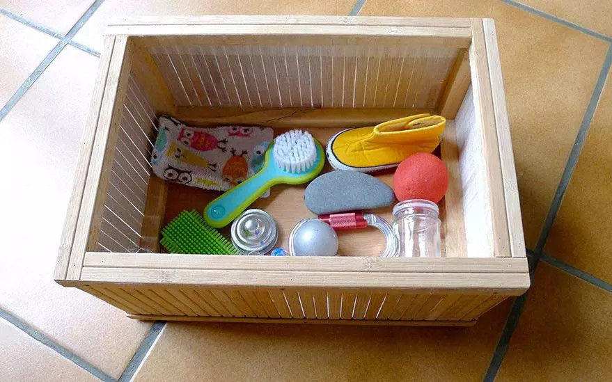 Une bo�te en bois contenant divers articles de puericulture, notamment une brosse verte pour bebe, une chaussure jaune, une balle rouge et un tissu a motifs representant des chouettes. La bo�te est posee sur un sol carrele et les articles sont soigneusement ranges. Le decor suggere un environnement domestique axe sur les articles essentiels pour bebe.