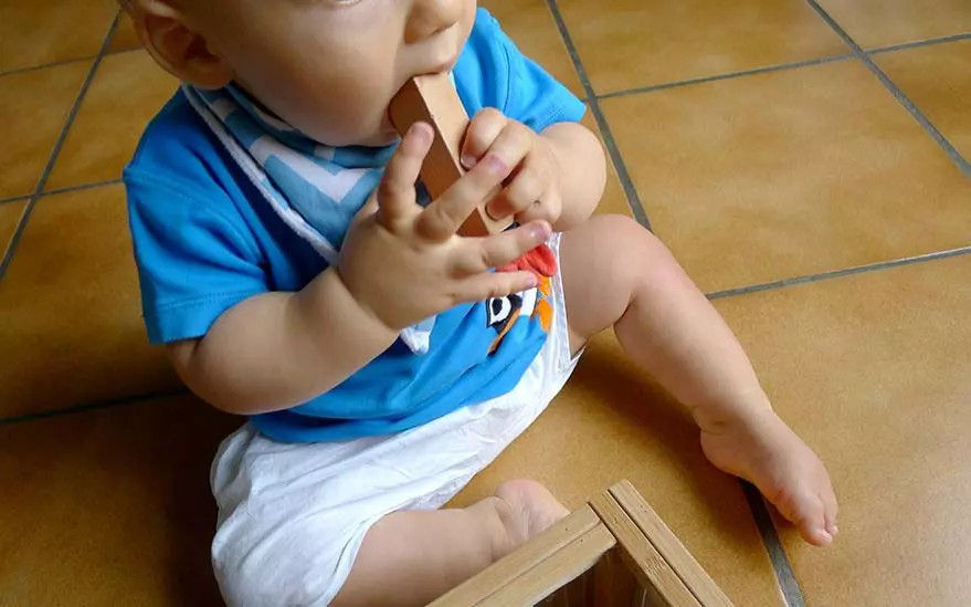 Un jeune bebe est assis sur un sol carrele, vetu d'une chemise bleue et d'un short blanc, et m�chouille un jouet en bois. Le decor semble etre un interieur avec un carrelage aux tons chauds. Le bebe explore de maniere ludique, faisant preuve de curiosite et de comportements propres au developpement precoce.