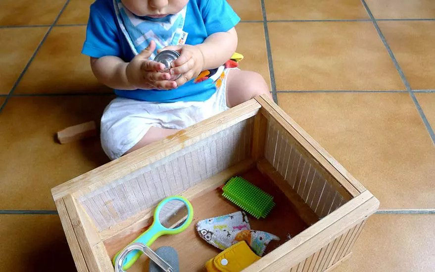 Un jeune enfant est assis sur un sol carrele et explore les objets contenus dans une bo�te en bois. Le bebe porte une chemise bleue et un short blanc, et tient un petit objet metallique. La bo�te contient des objets colores, notamment un peigne vert, une loupe et des morceaux de tissu.