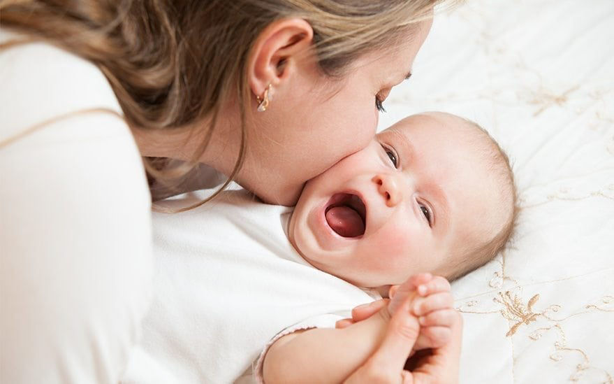 Une femme embrasse doucement un bebe allonge sur un lit recouvert d'un couvre-lit brode. Le decor est intime et chaleureux, mettant l'accent sur l'attention et la tendresse maternelles. La literie presente de delicats motifs floraux, ajoutant une touche esthetique douce et confortable a la scene.