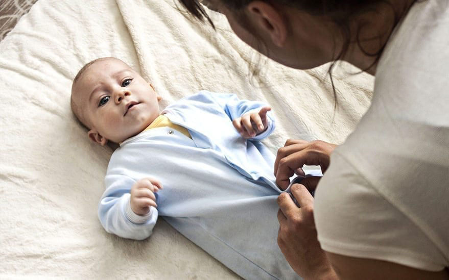 Un bebe est allonge sur une surface douce de couleur neutre, vetu d'une grenouillere bleu clair. On voit un adulte ajuster la tenue, en se concentrant sur les boutons. Le decor semble etre interieur, avec un eclairage naturel et une atmosphere chaleureuse.