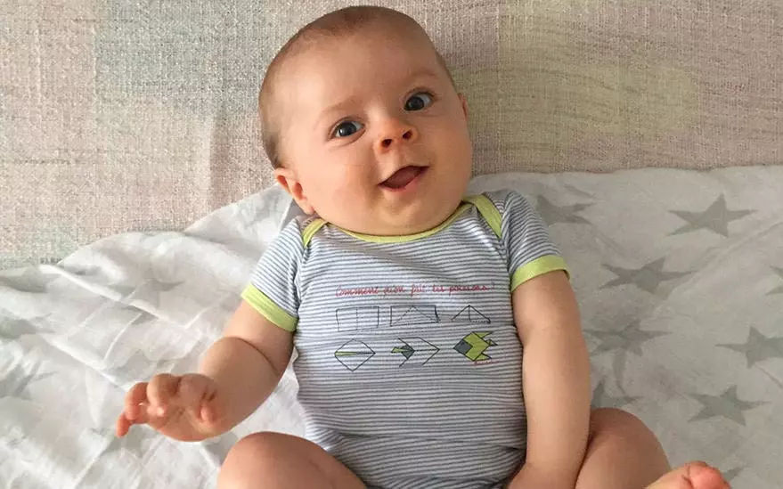 Un bebe vetu d'une grenouillere rayee bleu clair et blanc avec des formes geometriques et des inscriptions est assis sur une couverture douce a motifs d'etoiles. La grenouillere presente un melange de fleches et de formes dans des tons verts et gris. Le decor semble etre interieur, avec un arriere-plan aux tons neutres.