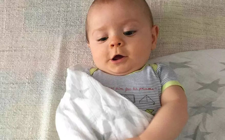 Un bebe est allonge sur une surface moelleuse, partiellement enveloppe dans une couverture blanche a motifs d'etoiles subtils. Le bebe porte une grenouillere rayee avec des motifs geometriques et des accents verts sur les manches. Le decor semble calme et confortable, avec un arriere-plan aux tons neutres.