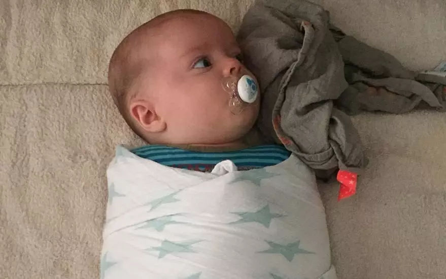 Un bebe enveloppe dans une couverture blanche a motifs d'etoiles vertes est allonge sur une surface beige douce. a proximite, un tissu gris avec une etiquette rouge est visible, ajoutant de la texture a la scene. Le decor suggere un environnement calme et confortable, adapte aux soins des nourrissons.