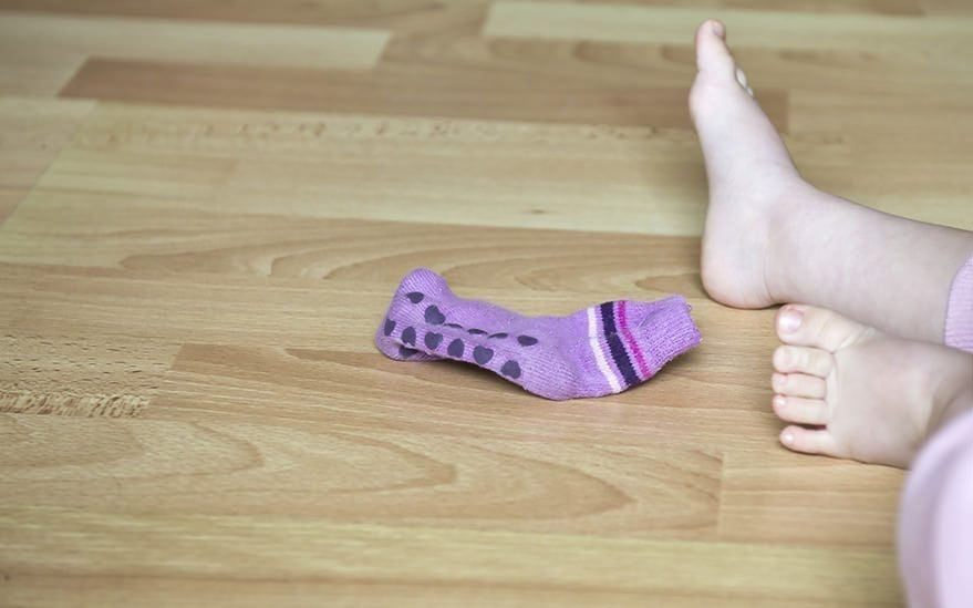 On aper�oit les pieds nus d'un enfant sur un parquet en bois, a c�te d'une chaussette violette a pois et rayures. Le decor suggere un environnement interieur decontracte. Le parquet presente une finition claire et naturelle.