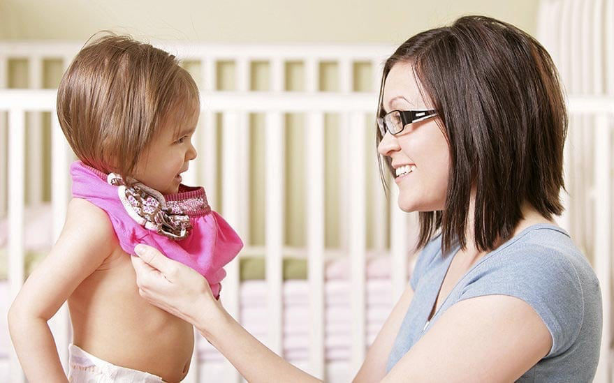 Une femme aide un enfant en bas �ge a mettre une echarpe rose dans une creche. L'arriere-plan presente un lit blanc et des tons pastel doux, creant une atmosphere chaleureuse et reconfortante. L'echarpe comporte des details decoratifs, ajoutant une touche ludique a la scene.