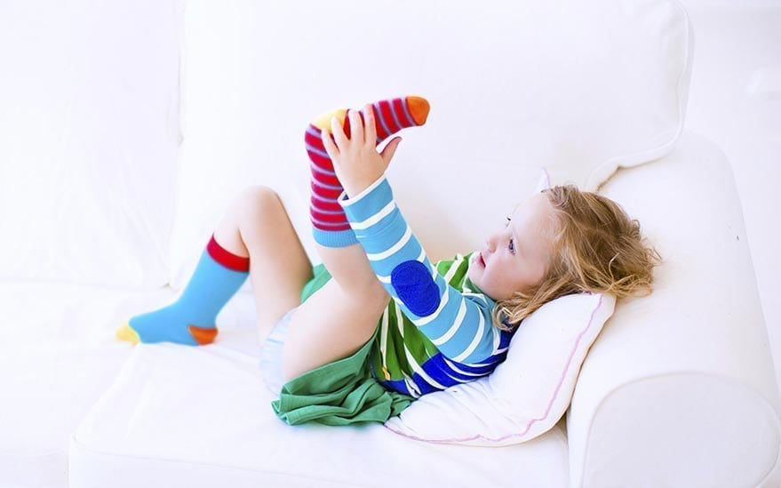 Un jeune enfant est allonge sur un canape blanc, etirant ses jambes de maniere ludique tout en portant des chaussettes rayees de couleurs vives. Le decor est un environnement interieur confortable avec un eclairage tamise et une decoration minimaliste. L'enfant est vetu d'une tenue aux couleurs vives, avec des rayures et des couleurs unies, creant une palette visuelle joyeuse et ludique.