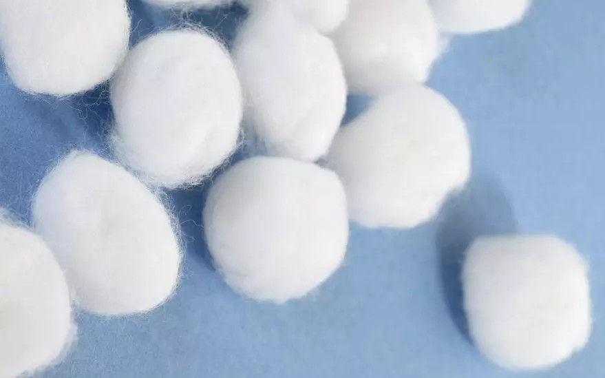 Gros plan sur plusieurs boules de coton blanc disposees sur une surface bleue lisse. L'image met en valeur la texture douce et l'aspect propre des boules de coton. Le decor est minimaliste, soulignant la simplicite et l'utilite du produit.