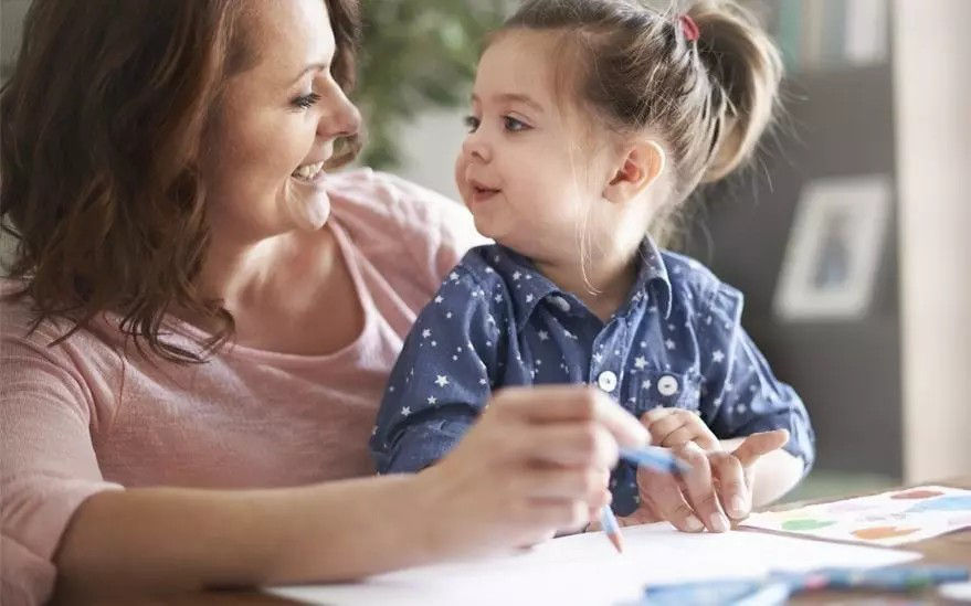 Une femme et une jeune fille sont assises a une table et s'adonnent a une activite de dessin. L'enfant tient un crayon bleu et des dessins colores sont visibles sur la table. Le decor semble etre un environnement interieur confortable avec un eclairage tamise et des elements d'arriere-plan flous.