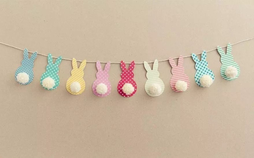 Une guirlande decorative composee de decoupes en forme de lapins aux couleurs pastel et a motifs a pois. Chaque lapin a une queue blanche duveteuse et est enfile sur un cordon fin. Le fond beige neutre met en valeur le design ludique et festif, ideal pour P�ques ou les celebrations printanieres.