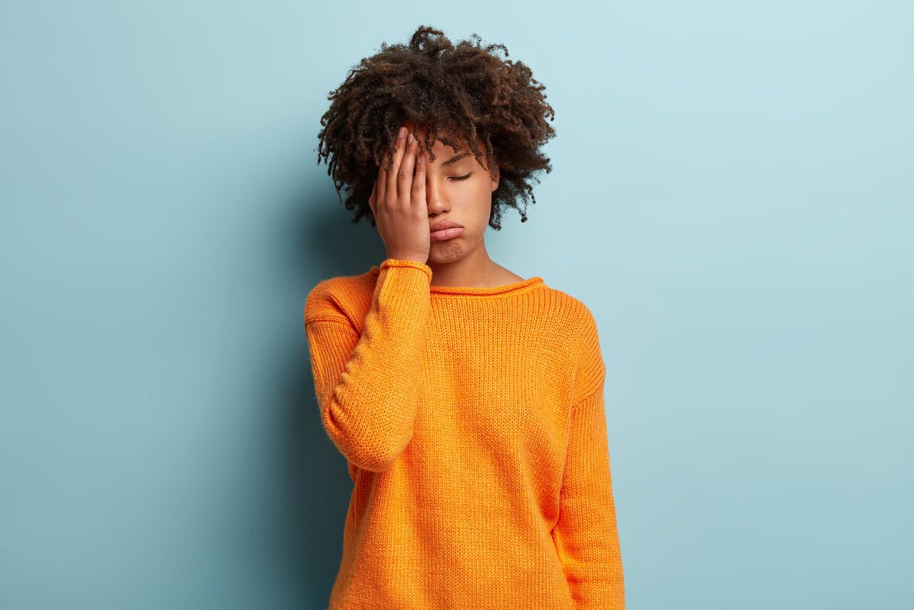 Photo of tired dark skinned female covers face, closes eyes, feels fatigue, needs good rest, dressed in orange jumper, has sleepy expression, isolated over blue background. Weariness and people