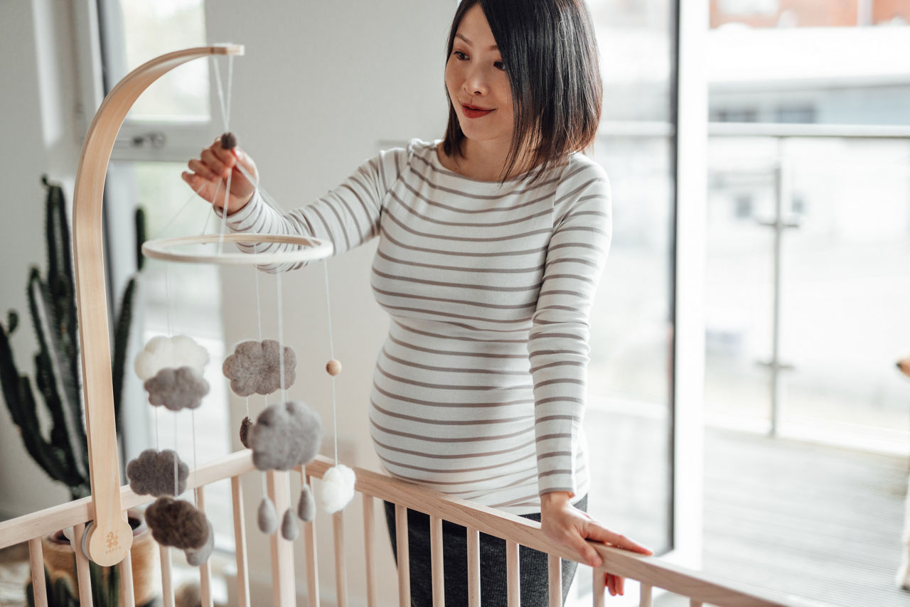 Beautiful Asian pregnant woman hanging a baby mobile onto a wooden crib. Feeling excited to prepare for the new baby.