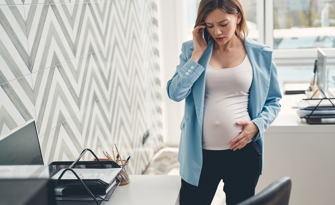 Worried expectant lady in blue jacket looking at belly while having phone conversation at work