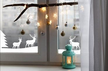 Une vitrine chaleureuse sur le theme des fetes, avec des decorations suspendues et des lumieres en forme d'etoiles sur une branche. Des autocollants blancs representant des rennes et des arbres ornent la vitre, creant une scene hivernale. Une lanterne turquoise avec une bougie allumee ajoute de la chaleur a l'ensemble.
