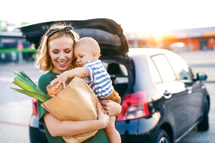 Beautiful young mother with her little baby son in front of a supermarket, holding paper shopping bag. Woman with a boy standing by the car.