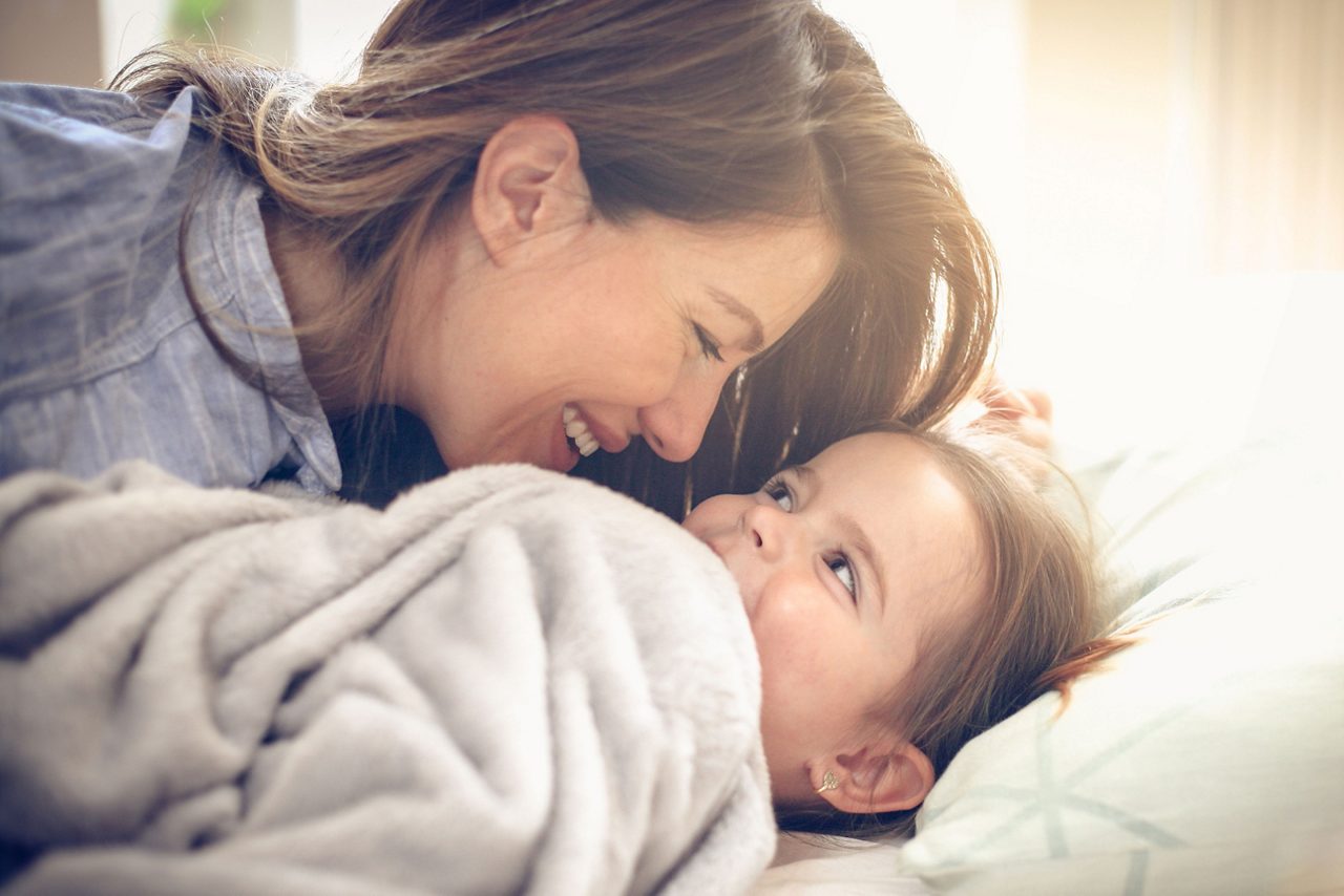 Mutter kuschelt mit zugedecktem Kleinkind im Bett rechts