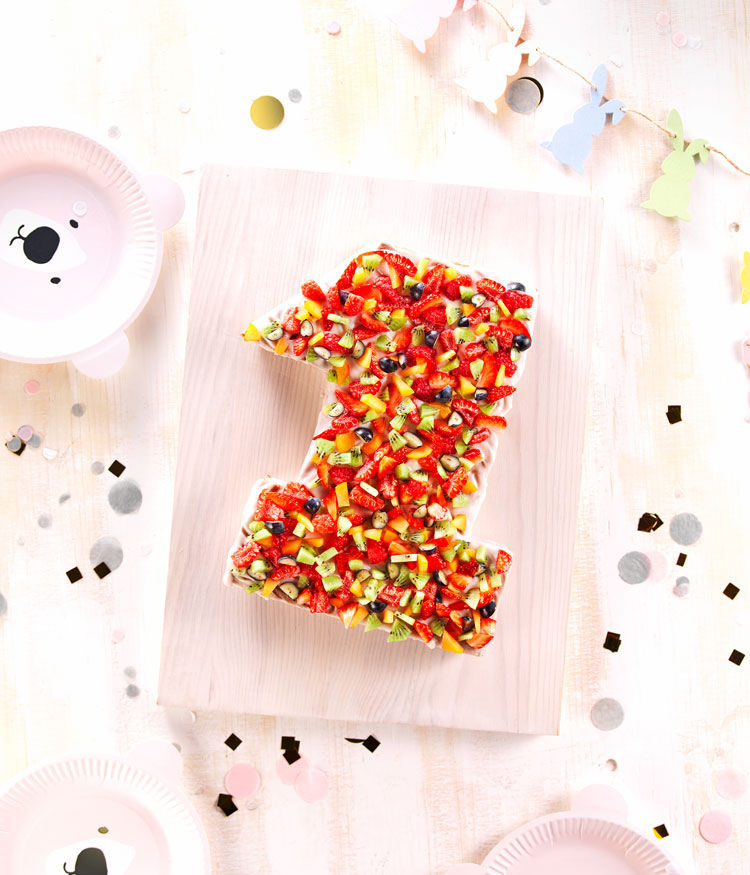 Un g�teau colore en forme de chiffre un, garni d'un assortiment de fruits frais, notamment des fraises, des kiwis et des myrtilles. Le g�teau est presente sur une planche en bois clair, entoure de decorations de fete telles que des confettis et des assiettes sur le theme des animaux. Le decor suggere une celebration joyeuse, probablement pour un premier anniversaire.
