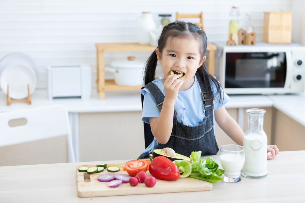 cute asian little child girl eating slice a cucumber with different vegetables in the kitchen 