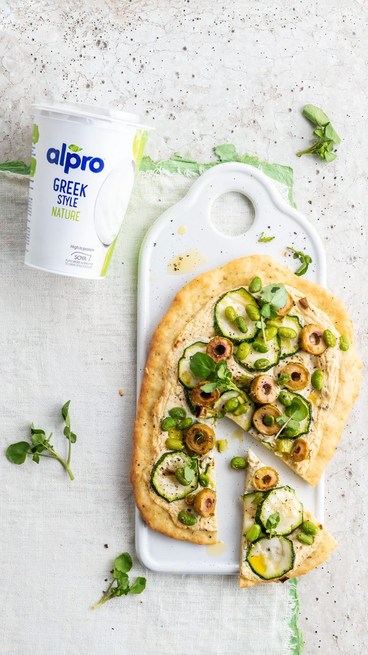 D69842DB-8B6C-4D7A-A03BA2EFCDCA466F<br>A_GFY_NA_Master_Recipe_Image_PizzaBianca_9x16__PBAQ_NA_Soy_NA_Pot_500g_HR_vertical-475.tif<br>Pizza Bianca 
Go green, with this veg-packed pizza made with Alpro Plain soya alternative to Greek style yogurt
"The dough:
400 g self-raising flour + extra for shaping
1 tbsp Italian seasoning + extra to finish off
1 tsp salt
300g Alpro soya alternative to Greek style yogurt plain

The topping:
1 tbsp fresh sage
3 spring onion
½ zucchini
12 green olives, pitted
200g Alpro Plain soya alternative to Greek style yogurt (50ml per pizza)
2 tbsp nutritional yeast flakes
A handful of  frozen peas or edamame beans, defrosted 
A handful of watercress, to serve"
"1. Start by pre-heating your oven to 200°C and cover a large baking tray with a baking sheet. Then make the dough. In a large bowl, whisk together the flour, salt and italian seasoning. (if you don’t have pre-mixed seasoning, make your own by grinding a mix of dried basil, oregano, rosemary, thyme, sage and coriander and blending with a little garlic powder.) Add the Alpro Plain soya alternative to Greek style yogurt, and stir together well using a rubber spatula until you have a smooth dough.
2. Divide the dough into four and flatten each piece with your hands to make a pizza shape and arrange them on your baking tray. 
3. Now make the sauce. Finely chop the sage and add it to the Alpro Plain soya alternative to Greek style yogurt along with the nutritional yeast flakes. Season with salt and pepper to taste. Then on to the topping. Chop the spring onions, slice the zucchini and halve the olives. 
4. Now the final assembly. Cover the pizza doughs with the sauce and finish with the spring onion, zucchini, green olives and peas. Sprinkle with olive oil, season with salt, pepper and scatter on a few extra Italian herbs. Bake in the hot oven for 15-20 minutes and serve with watercress and any sauce that’s left over, if you like. 

Tip: If you want to cook this on a BBQ, pre-bake the pizza doughs until they start to brown. (5-6 min) then take them off, add the sauce and toppings and put back on the BBQ until the veggies are nice an soft."
lactose free, veggie, vegan
/pizza-bianca
(Recipe type: MAINS (Prep time: 25 min) (baking time: 15-20 min) (servings: +/- 4 persons) (season: all year) (pool: 1) NOT TO BE TRANSLATED
/
/
To make it extra green, add some finely chopped spinach to the sauce
Gluten free? No problem - use gluten-free self-raising flour.
Alpro Plain soya alternative to Greek style yogurt
Alpro, Alpro recipe, pizza, bianca, greek, flatbread, green, veggie, vegan, BBQ, summer