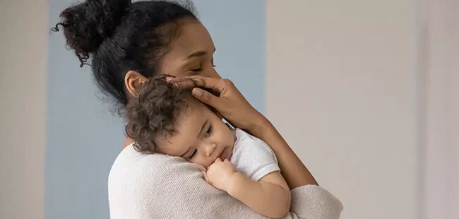 A woman is seen holding a baby in a warm and intimate indoor setting. The scene features soft, neutral tones with a light blue and beige background, creating a calm and nurturing atmosphere. The focus is on the bond between the woman and the child, emphasizing care and tenderness.
