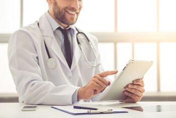 Cropped image of handsome medical doctor in white coat using a digital tablet and smiling while working in his office