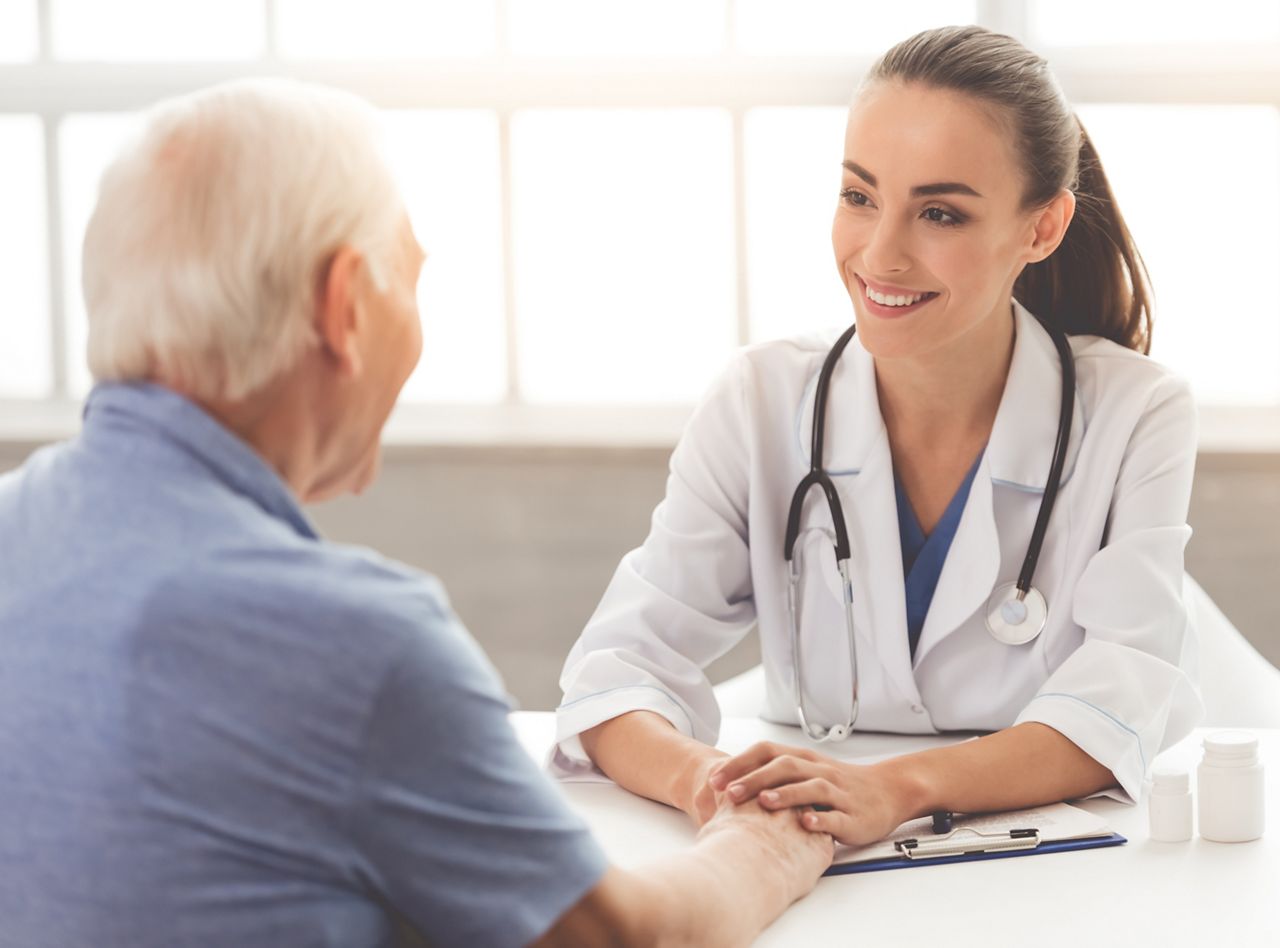 Beautiful female doctor in white medical coat is consulting her patient, holding his hand and smiling while sitting in office