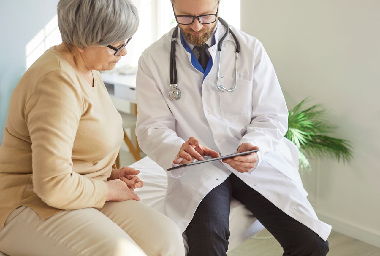 Doctor or nurse conducts medical consultation for elderly patient in hospital office, using tablet pc for care in a professional setting with focus on communication and healthcare support.