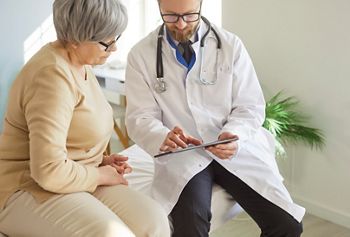 Doctor or nurse conducts medical consultation for elderly patient in hospital office, using tablet pc for care in a professional setting with focus on communication and healthcare support.