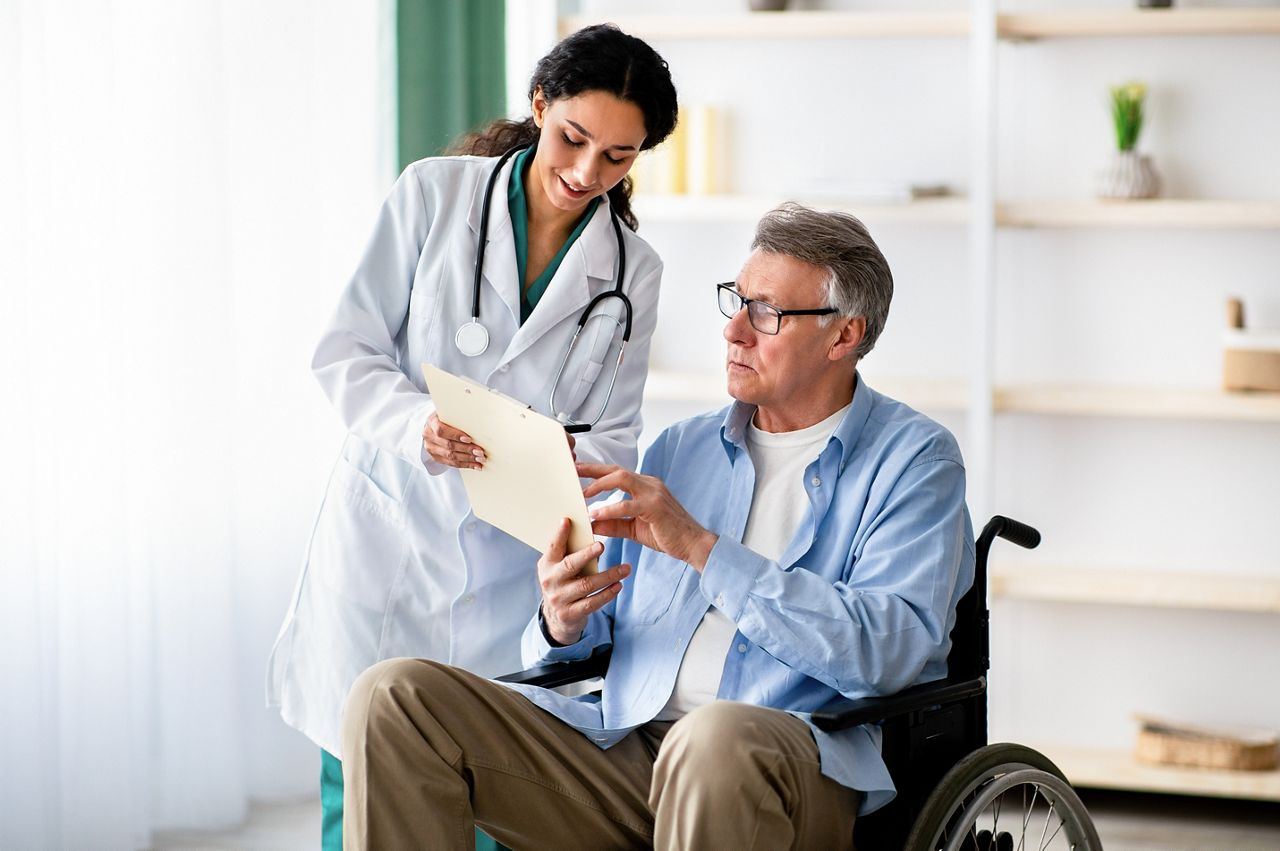 Young nurse offering handicapped elderly man in wheelchair to sign insurance contract at retirement home. Disabled older male putting signature under medical document, indoors