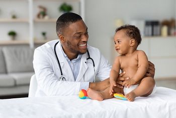 Child's Healthcare Concept. Portrait Of Smiling Black Doctor Making Check Up For Little Infant Baby Boy, Cute Toddler Child In Diaper Having Appointment At Pediatrician's Office, Free Space