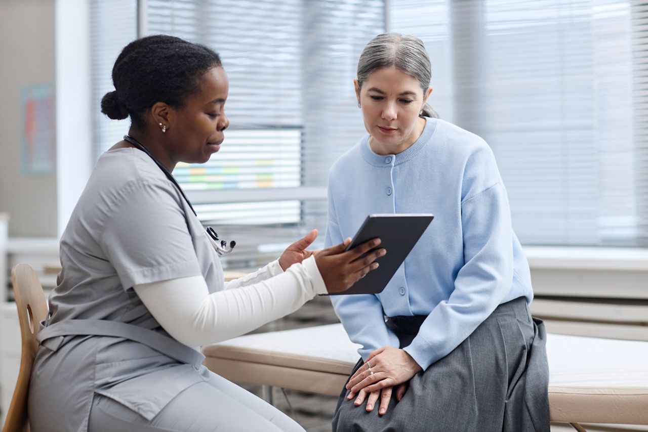 Black young adult woman nurse showing digital tablet to Caucasian middle aged woman patient during medical consultation in clinic examination room, both focused on screen