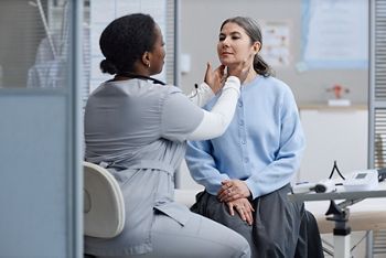 Black woman doctor examining neck of middle aged Caucasian woman during medical checkup in clinic, both women focused on health assessment, medical equipment visible on table nearby