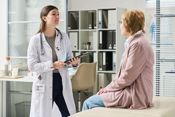 Young female doctor holding digital tablet speaking with senior woman sitting on examination table in medical office during consultation