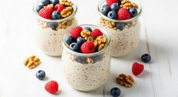 Creamy overnight oats in clear glass jars, topped with fresh blueberries, red raspberries, and crunchy walnuts on a bright wooden surface, vegetarian, jars, food