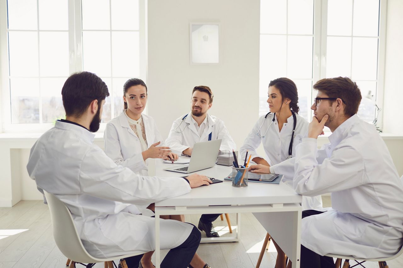 Group of practicing doctors in a meeting discuss the diagnosis of a patient sitting at desk in a clinic office. Confident doctors at the conference talking.
