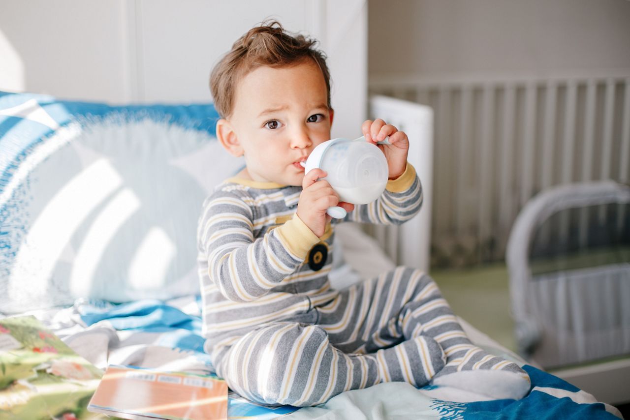 Cute adorable Caucasian kid boy sitting on bed drinking milk from kids bottle. Healthy eating drinking for little children. Supplementary food for growing babies. Candid real authentic moment.