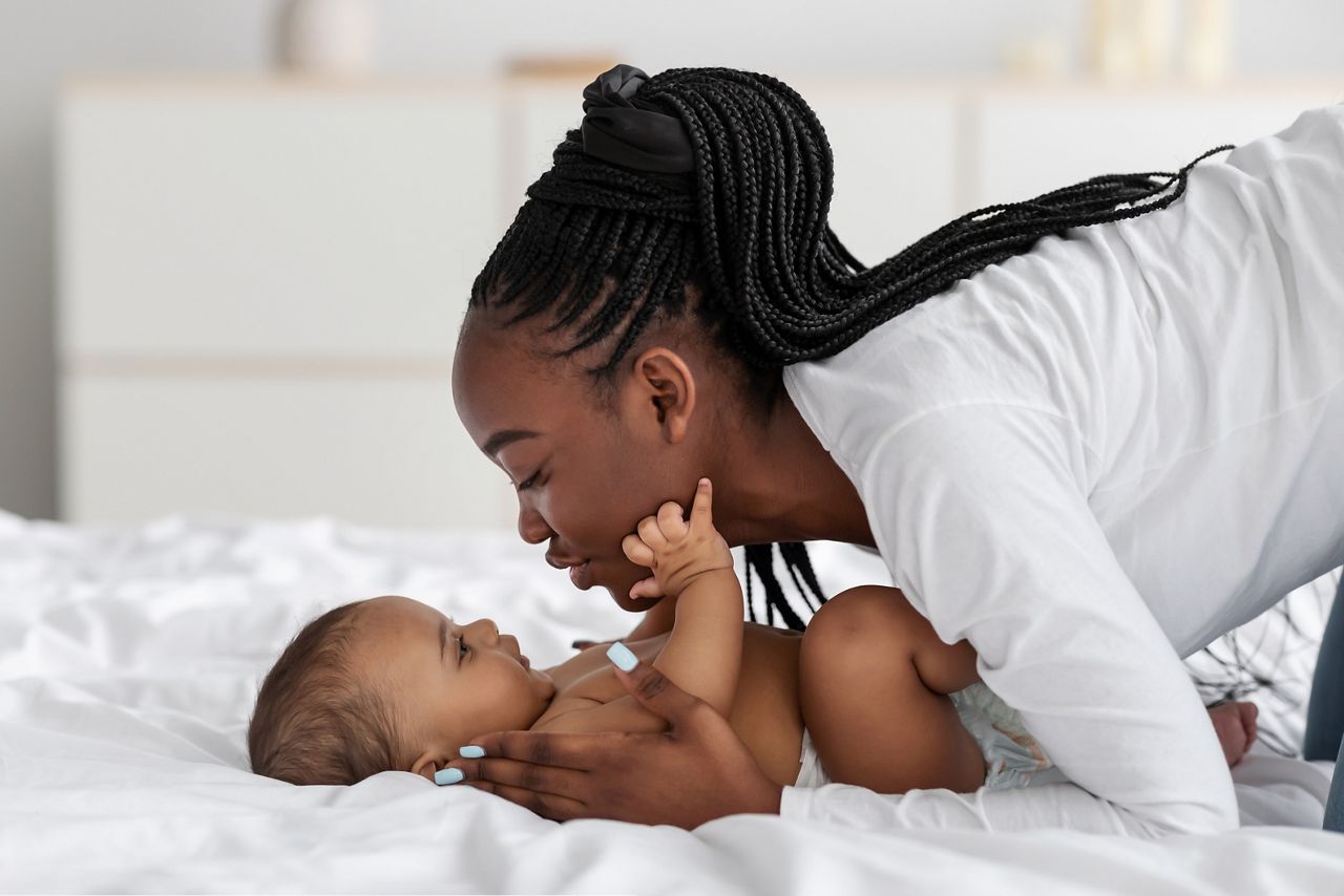 Happy Motherhood Concept. Closeup profile portrait of cheerful young black woman playing with her adorable small baby who lying in bed on white bedsheets, kissing sweet infant child