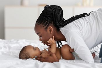 Happy Motherhood Concept. Closeup profile portrait of cheerful young black woman playing with her adorable small baby who lying in bed on white bedsheets, kissing sweet infant child