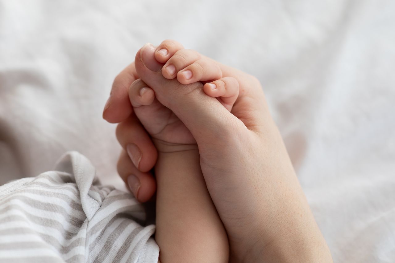 Mother's Protection. Closeup Shot Of Unrecognizable Mom Holding Tiny Hand Of Her Newborn Baby While They Relaxing Together On Bed At Home, Enjoying Bonding With Infant Child, Cropped Image