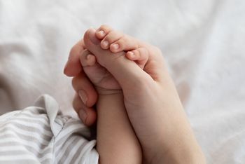 Mother's Protection. Closeup Shot Of Unrecognizable Mom Holding Tiny Hand Of Her Newborn Baby While They Relaxing Together On Bed At Home, Enjoying Bonding With Infant Child, Cropped Image