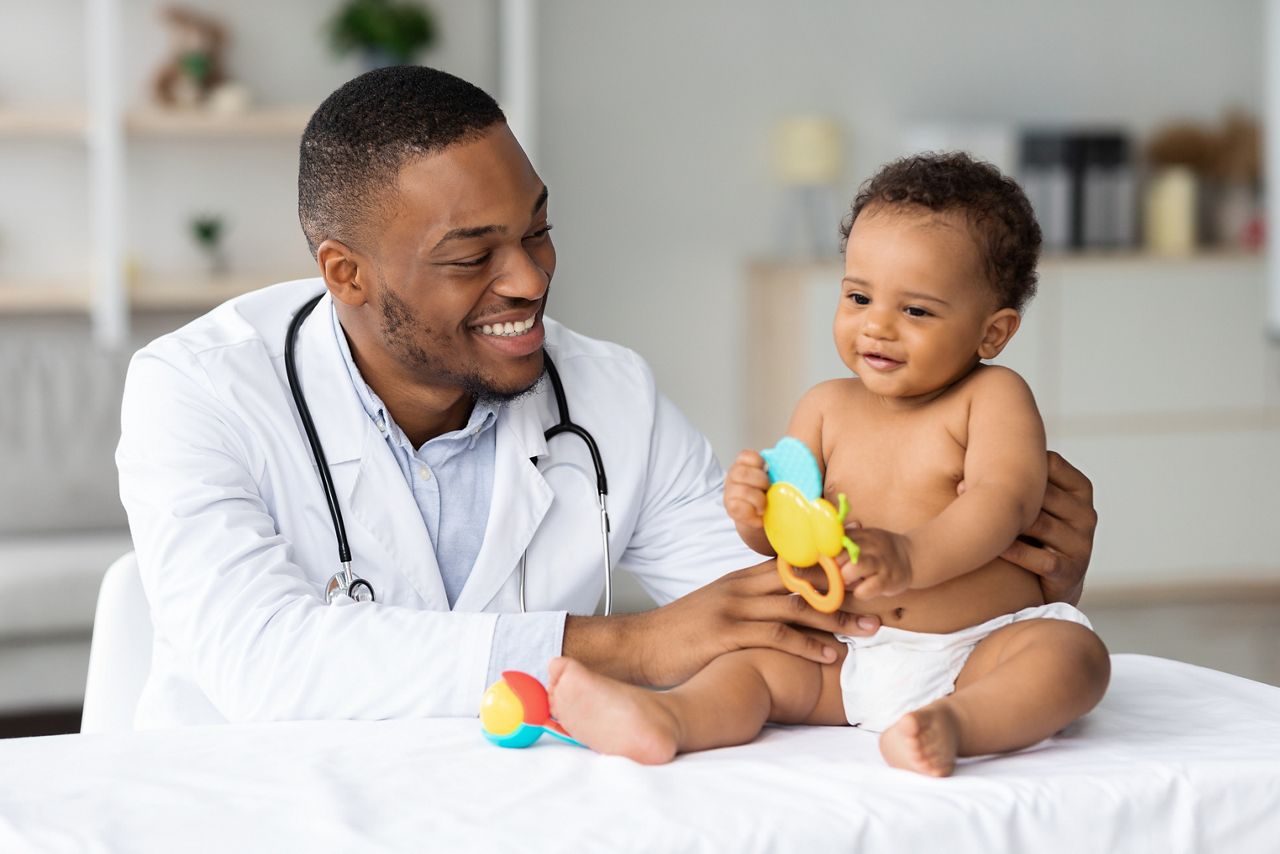 Portrait Of Smiling Black Doctor Making Medical Check Up For Little Baby Patient In Clinic, Handsome Young African American Pediatrist Looking At Adorable Infant Child In Diaper, Closeup Shot